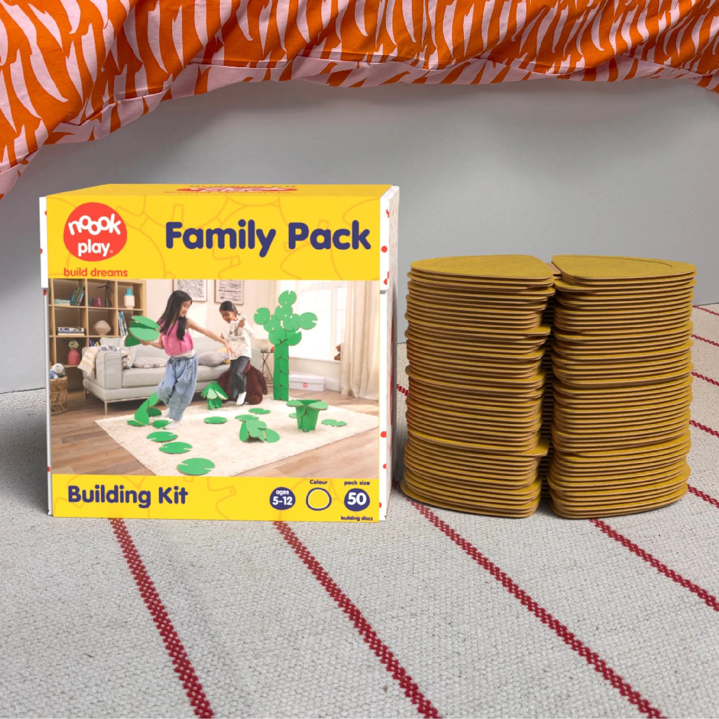 The noook play® Family Pack, 50 Pieces, in yellow, shown at the foot of a child bed, on a stripy rug