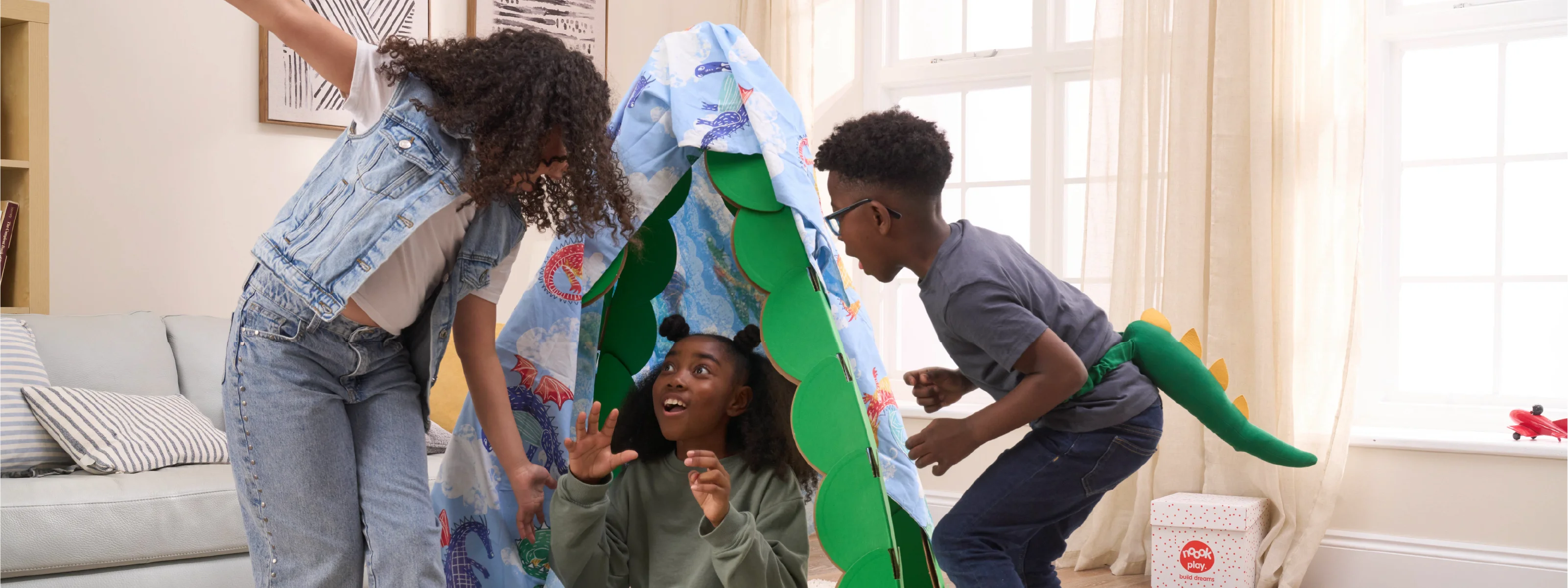 Three kids are playing in a living room, pretending to find dragons in a lair made out a noook play® building kit. The noook® den supports a sheet with dragons on.
