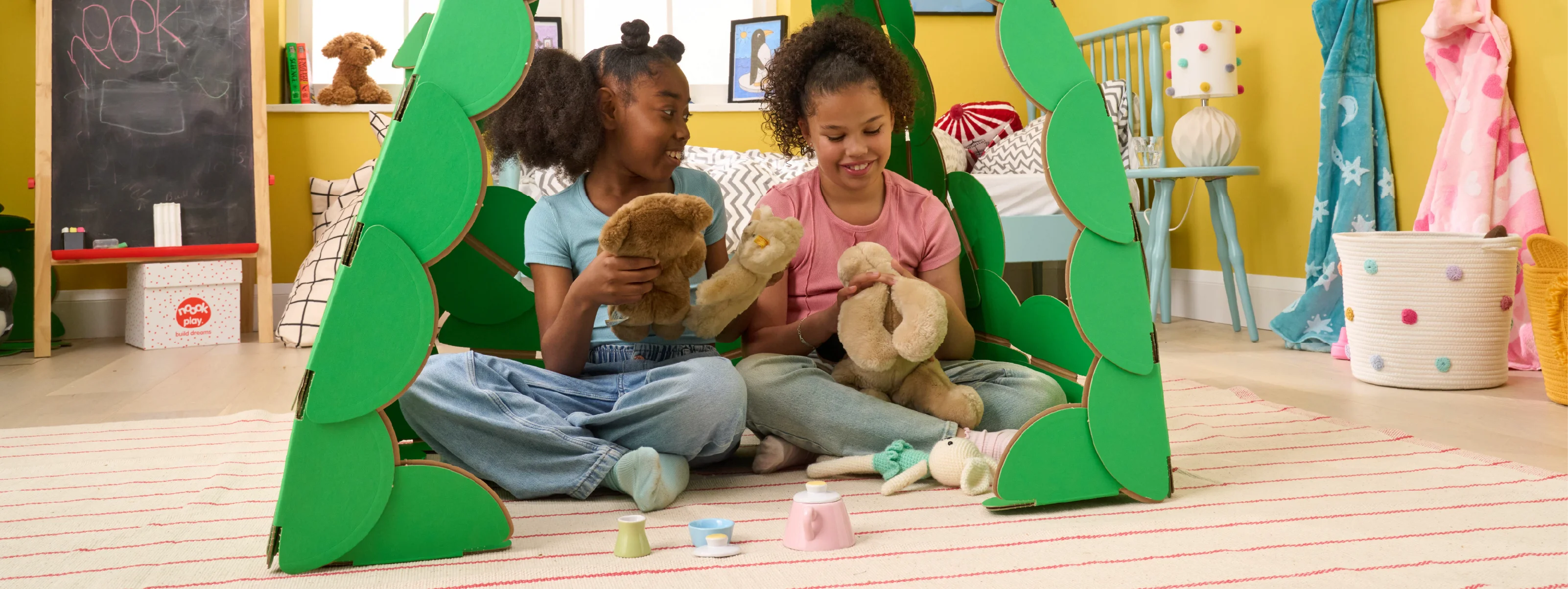 Two girls are playing in a bedroom with their teddies and a tea set, in a den made out of a noook play® building kit.